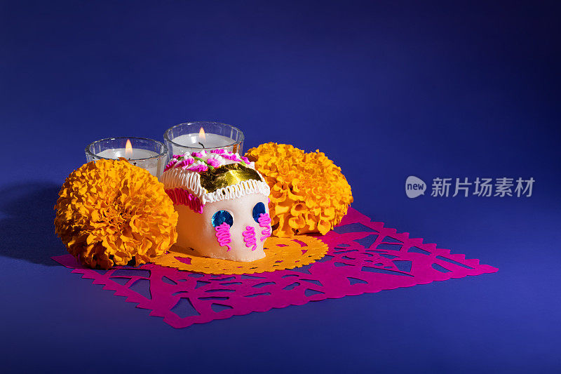 死者日装饰的糖头骨(calavera)祭奠蜡烛和金盏花在粉红色的picado在深蓝色背景，Dia de Muertos的概念